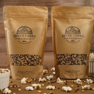 Puede incluir: Dos bolsas de papel marrón de granos de maíz para palomitas de maíz. La bolsa de la izquierda está etiquetada como "Autumn Medley" y la bolsa de la derecha está etiquetada como "Purple Popcorn". Ambas bolsas son de Seven Forks Farm, Family Grown Popcorn.