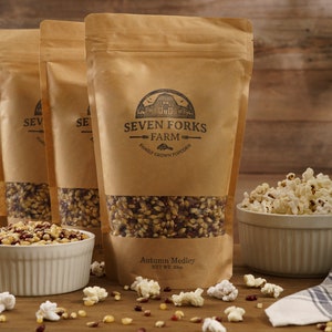 Puede incluir: Tres bolsas de papel marrón llenas de granos de palomitas de maíz. La bolsa del centro tiene el texto "Seven Forks Farm Family Grown Popcorn" y "Autumn Medley 10 oz. WT 283g". Un tazón de palomitas de maíz se encuentra sobre una mesa de madera.
