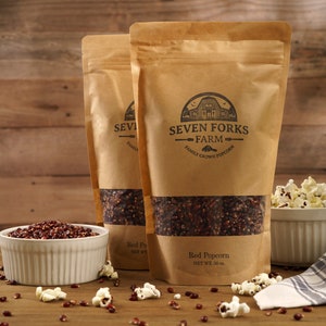 Op de afbeelding: Twee bruine papieren zakken gevuld met rode popcornkorrels. De zakken hebben een label met de tekst "Seven Forks Farm - Family Grown Popcorn". Er staat een kom met gepopte rode popcorn en een kom met rode popcornkorrels op een houten tafel.