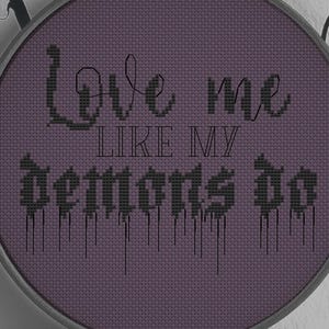 May include: A circular embroidery hoop containing purple fabric with black cross-stitched text that reads Love me LIKE MY demons do. The word demons features a dripping paint effect. An orange bird logo with the text BYMEGC is at the bottom.