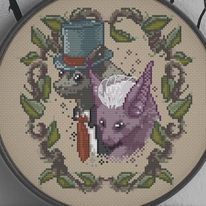 Puede incluir: Un bordado de punto de cruz circular en un aro de madera. El diseño presenta dos criaturas estilizadas, una con sombrero de copa y la otra con pelaje morado, enmarcadas por un borde de hojas verdes. El texto "By Meg C" está en la parte inferior.