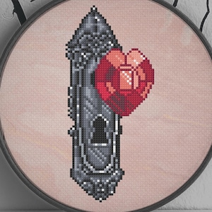 Op de afbeelding: Kruissteekkunst in een ronde lijst met een gedetailleerd, gepixeld ontwerp van een gotische deurklink en een rood hart. Het kunstwerk is geplaatst tegen een lichtroze achtergrond, met de tekst "By Meg C" onderaan. Decoratieve takken omlijsten de bovenkant.