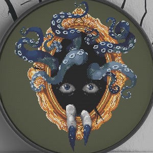 May include: A round, dark green cross-stitch design featuring a gold-framed mirror with a black void and blue octopus tentacles. The design includes two eyes, two long fingernails, and the text "By Meg C" with an orange raven logo.