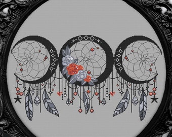 May include: A black and white cross stitch pattern of three crescent moons with dreamcatchers and feathers hanging from them. The moons are surrounded by small red berries and white flowers. The pattern is framed in a black ornate frame.
