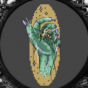 May include: Cross-stitch art in a black ornate frame. The artwork depicts a green hand with purple nails holding a wasp. The background is a dark gray oval with a gold-colored border. The frame has a crow on top with the text "By Meg C".