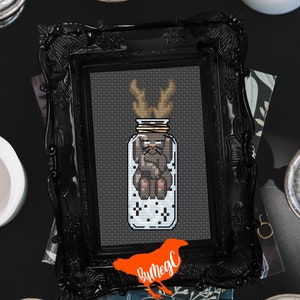 May include: A cross-stitch pattern of a deer with antlers inside a glass jar with a white background. The jar has a brown lid and a brown twine bow. The deer is brown and has a white nose and belly. The background is gray.