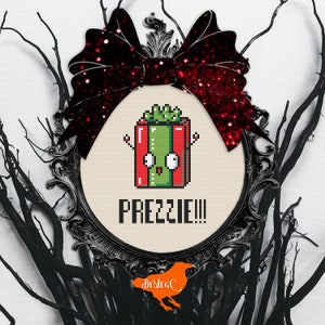 Puede incluir: Un diseño de punto de cruz de una caja de regalo de dibujos animados con una cara verde y la palabra "PREZZIE!!!" en negro. El diseño está enmarcado en un marco ovalado negro adornado con un lazo rojo brillante. Ramas negras rodean el marco.