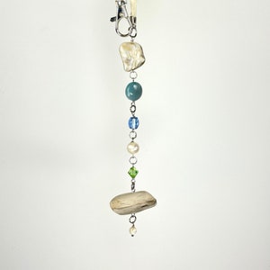 May include: A white cord with a silver clasp and a beaded charm. The charm features a white shell, blue and green beads, and a small piece of driftwood.