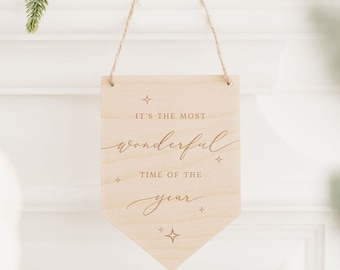 May include: Wooden sign with the text "It's the most wonderful time of the year" in a cursive font. The sign is hanging from a string and has a light wood finish.