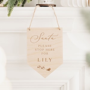 Op de afbeelding: Een houten bord met de tekst "Santa Please Stop Here For Lily" en een kleine illustratie van de slee van Sinterklaas met rendieren. Het bord hangt aan een touwtje en bevindt zich voor een witte schouw.