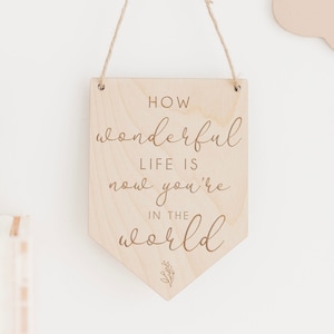 May include: Wooden sign with the text "How wonderful life is now you're in the world" in a cursive font. The sign is hanging from a string and has a small floral design at the bottom.