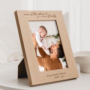May include: A light wood photo frame with a personalized inscription that reads "First Christmas as my Daddy" and "Love from Oliver 2024". The frame features a photo of a father holding his baby.