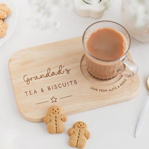May include: Wooden tea and biscuit serving board with the text "Grandad's TEA & BISCUITS". A glass mug of tea sits on the board, with two gingerbread men biscuits. A gold spoon and a plate of biscuits are also present.