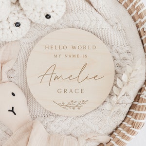 May include: A round wooden sign with the text "Hello World My Name Is Amelie Grace" and a small floral design.