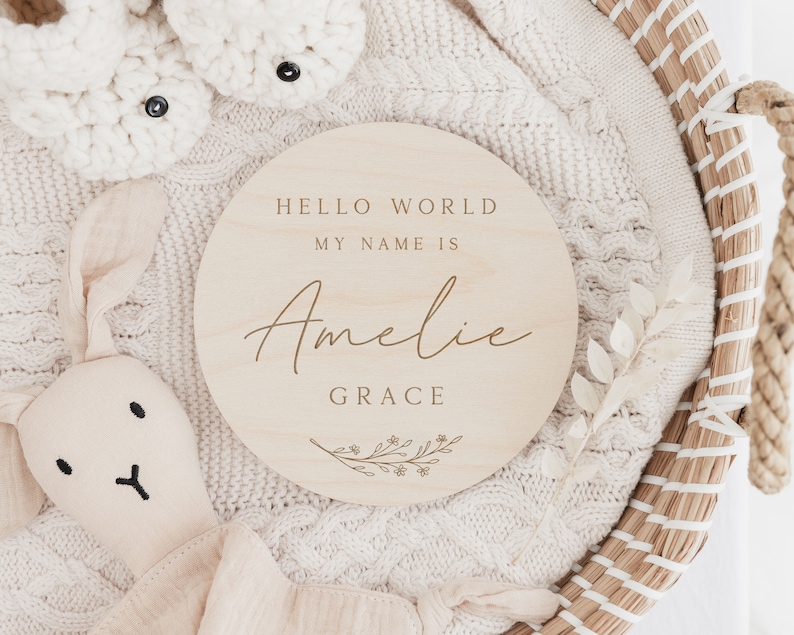 May include: A round wooden sign with the text "Hello World My Name Is Amelie Grace" and a small floral design.