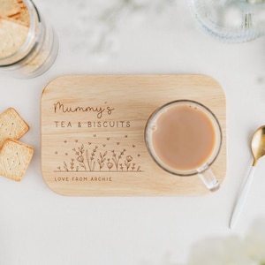 K&ouml;nnte beinhalten: Ein h&ouml;lzernes Tee- und Keks-Servierbrett mit dem Text "Mummy's TEA & BISCUITS" und einem floralen Design. Eine klare Glastasse mit Tee und ein goldener L&ouml;ffel befinden sich auf dem Brett. Kekse und ein Keksdose sind ebenfalls im Bild.
