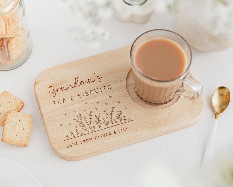 K&ouml;nnte beinhalten: Ein Holztablett f&uuml;r Tee und Kekse mit einer Glastasse Tee. Auf dem Tablett steht "Grandma's TEA & BISCUITS" und "LOVE FROM OLIVER & LILY". Ein goldener L&ouml;ffel und Kekse sind ebenfalls vorhanden.