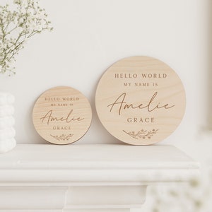 May include: Two round wooden signs with the text "Hello World My Name Is Amelie Grace" in a cursive font. The signs are on a white mantel.