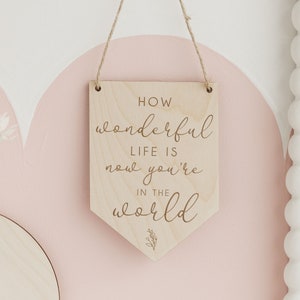 May include: Wooden sign with the text "How wonderful life is now you're in the world" in a cursive font. The sign is hanging from a string and has a light wood finish.