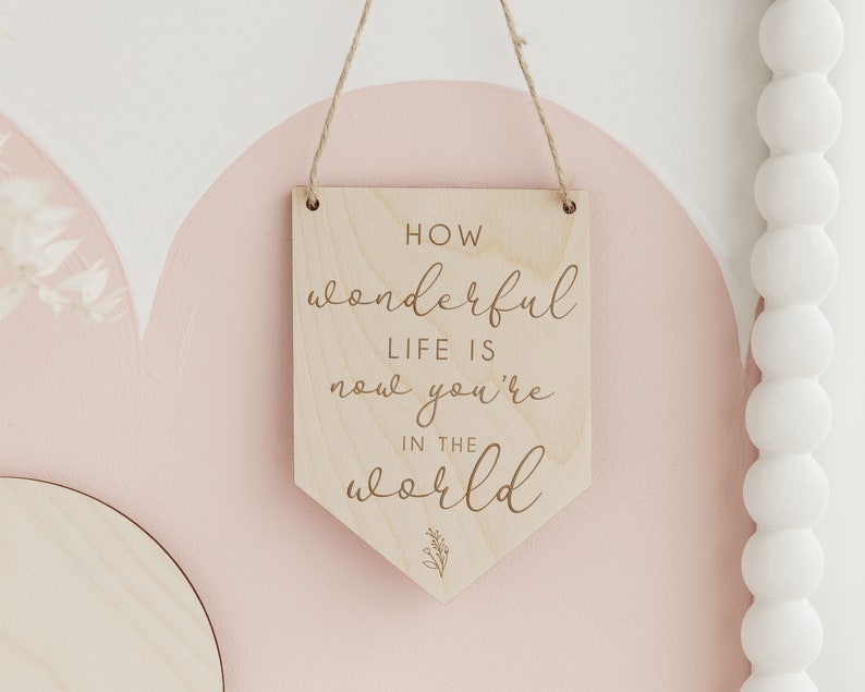 May include: Wooden sign with the text "How wonderful life is now you're in the world" in a cursive font. The sign is hanging from a string and has a light wood finish.