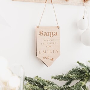 Op de afbeelding: Een houten bord met de tekst "Santa Please Stop Here For Emilia" en een kleine illustratie van een rendier dat een slee trekt. Het bord hangt aan een touwtje en is tegen een witte muur.