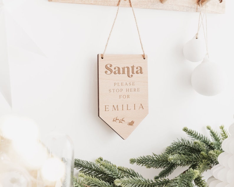 Op de afbeelding: Een houten bord met de tekst "Santa Please Stop Here For Emilia" en een kleine illustratie van een rendier dat een slee trekt. Het bord hangt aan een touwtje en is tegen een witte muur.