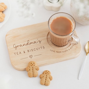 K&ouml;nnte beinhalten: Ein h&ouml;lzernes Tee- und Keks-Serviertablett mit dem Text "Grandma's TEA & BISCUITS" und einem Herzsymbol. Auf dem Tablett steht eine Glastasse Tee, zusammen mit zwei Lebkuchenm&auml;nnern und einem goldenen L&ouml;ffel.