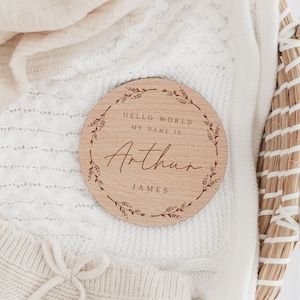 May include: A round wooden sign with the name "Arthur" engraved in a script font. The sign includes the text "HELLO WORLD MY NAME IS" and "JAMES" in a simple font. A decorative wreath of leaves surrounds the text.