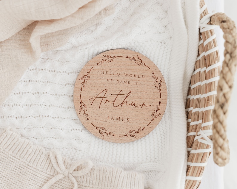 May include: A round wooden sign with the name "Arthur" engraved in a script font. The sign includes the text "HELLO WORLD MY NAME IS" and "JAMES" in a simple font. A decorative wreath of leaves surrounds the text.