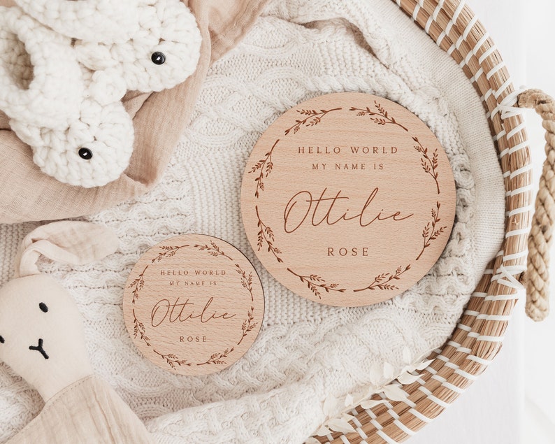 May include: Two wooden baby announcement signs with the text "HELLO WORLD MY NAME IS Ottilie ROSE" surrounded by a floral wreath. A white crocheted bunny and a white crocheted bunny head are also in the image.