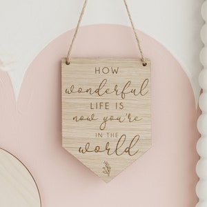 May include: Wooden sign with the text "How wonderful life is now you're in the world" in a cursive font. The sign is hanging from a string and has a small leaf design at the bottom.