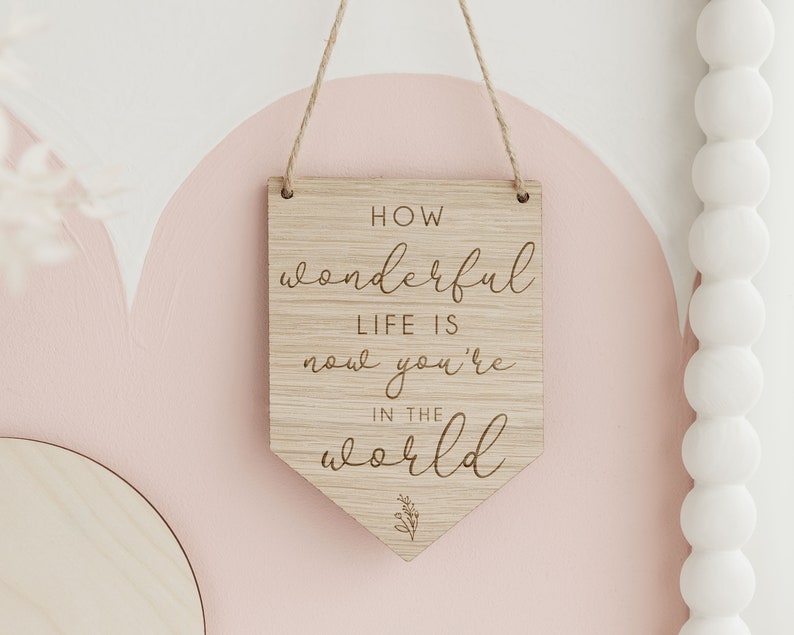 May include: Wooden sign with the text "How wonderful life is now you're in the world" in a cursive font. The sign is hanging from a string and has a small leaf design at the bottom.