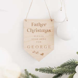 Op de afbeelding: Houten bord met de tekst "Kerstman Stop hier alstublieft voor George" en een klein beeld van de slee van de kerstman met rendieren.