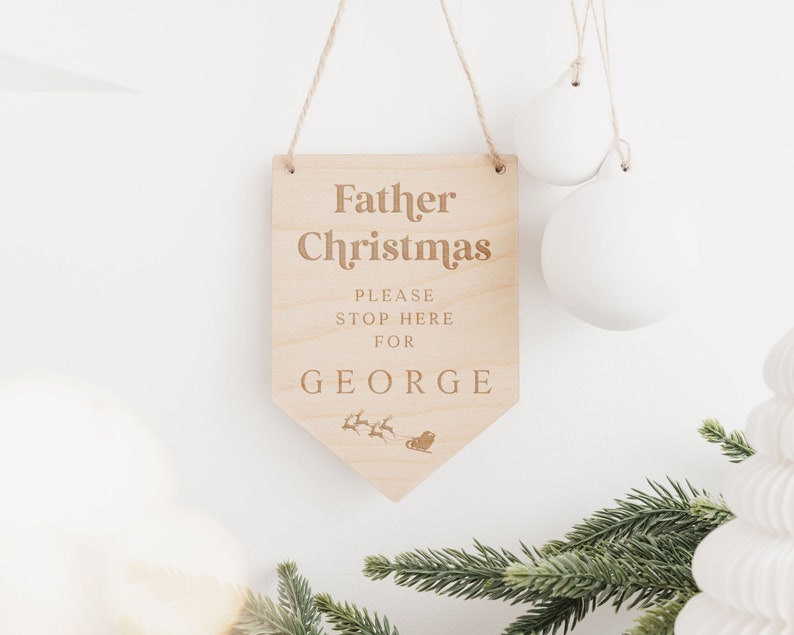 Op de afbeelding: Houten bord met de tekst "Kerstman Stop hier alstublieft voor George" en een klein beeld van de slee van de kerstman met rendieren.