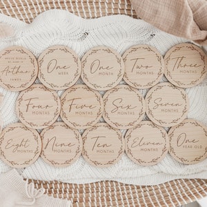 May include: A set of twelve wooden milestone discs with engraved text and leaf border designs. The discs mark stages from "One Week" to "One Year Old", with a disc that says "Hello World, My Name Is Arthur James".