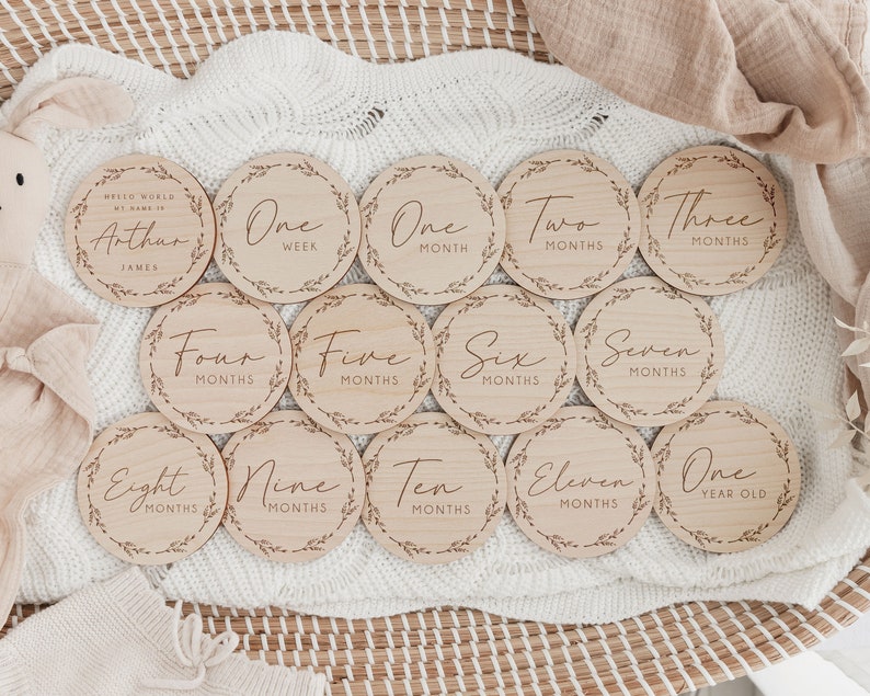 May include: A set of twelve wooden milestone discs with engraved text and leaf border designs. The discs mark stages from "One Week" to "One Year Old", with a disc that says "Hello World, My Name Is Arthur James".