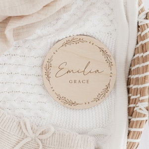 May include: A round wooden plaque with a floral design and the name "Emilia Grace" engraved in cursive.