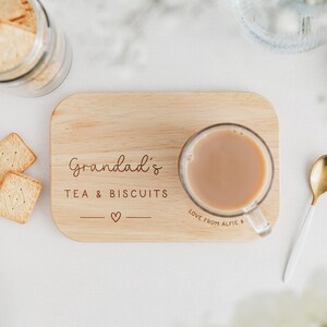 May include: A wooden serving tray with the words "Grandad's TEA & BISCUITS" and a heart symbol. A clear glass mug of tea sits on the tray, next to a gold and white spoon. A jar of biscuits and two biscuits are also in the image.