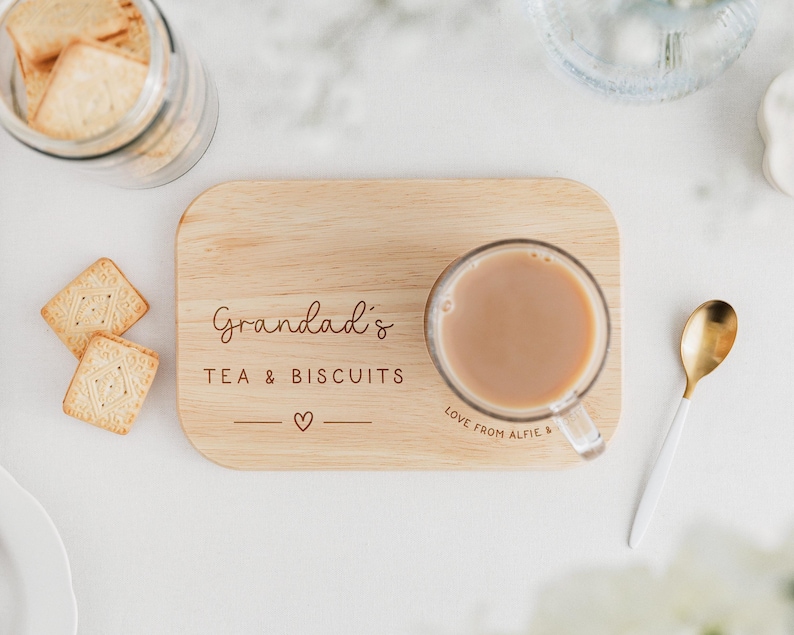May include: A wooden serving tray with the words "Grandad's TEA & BISCUITS" and a heart symbol. A clear glass mug of tea sits on the tray, next to a gold and white spoon. A jar of biscuits and two biscuits are also in the image.