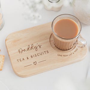 May include: A wooden tea and biscuit serving board with a clear glass mug of tea. The board is engraved with "Daddy's TEA & BISCUITS" and a heart symbol. A gold spoon and a jar of biscuits are also visible.