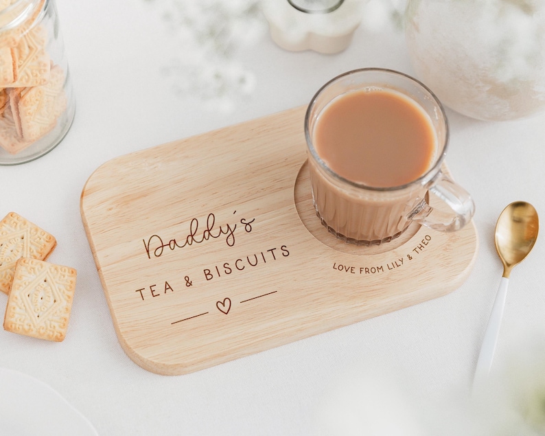 May include: A wooden tea and biscuit serving board with a clear glass mug of tea. The board is engraved with "Daddy's TEA & BISCUITS" and a heart symbol. A gold spoon and a jar of biscuits are also visible.