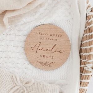May include: A round wooden sign with the text "Hello World My Name Is" and the name "Amelie Grace" in cursive script. The sign is decorated with a floral design.