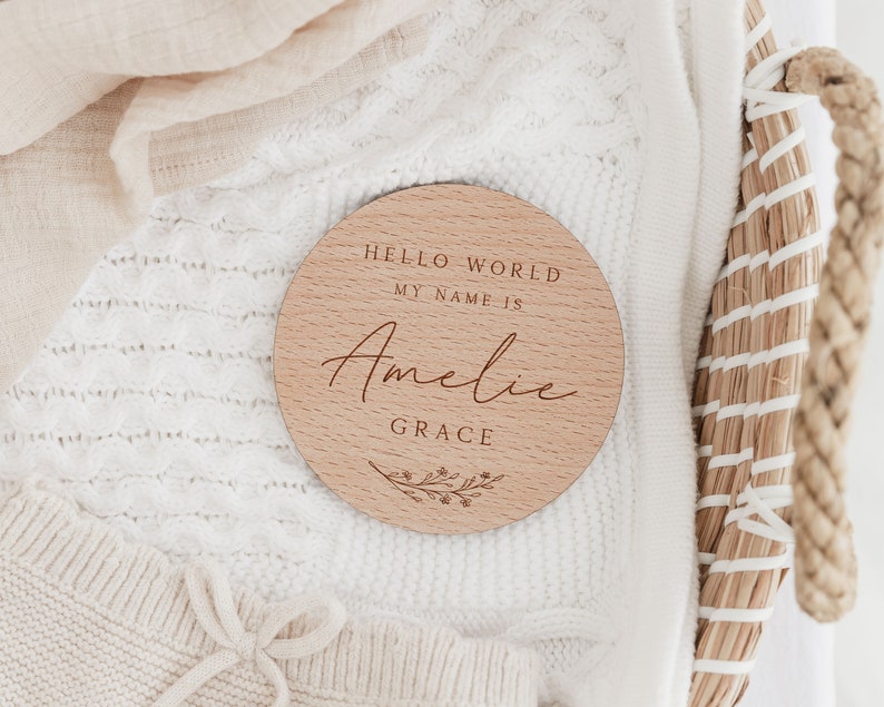 May include: A round wooden sign with the text "Hello World My Name Is" and the name "Amelie Grace" in cursive script. The sign is decorated with a floral design.