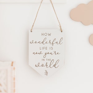 May include: White wooden sign with a brown inscription that reads "How wonderful life is now you're in the world". The sign is hanging from a string and has a small floral design at the bottom.