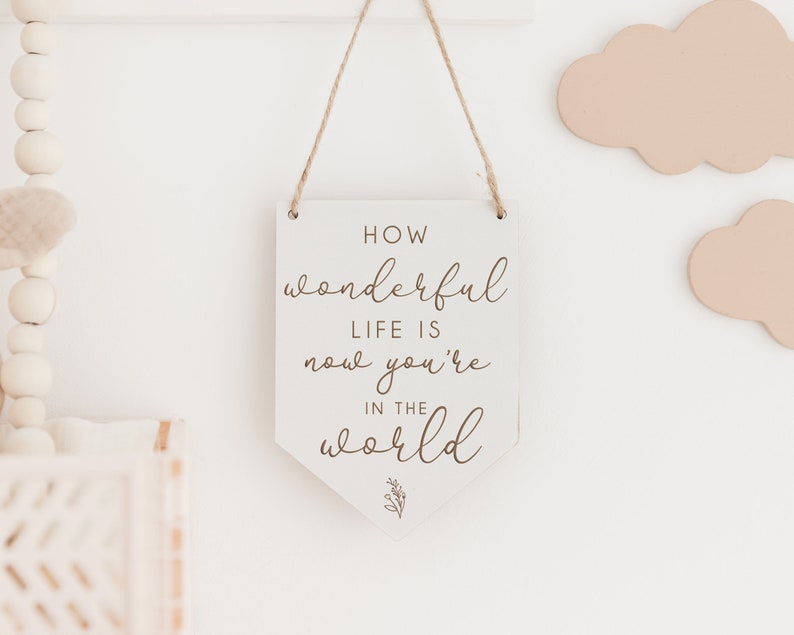May include: White wooden sign with a brown inscription that reads "How wonderful life is now you're in the world". The sign is hanging from a string and has a small floral design at the bottom.