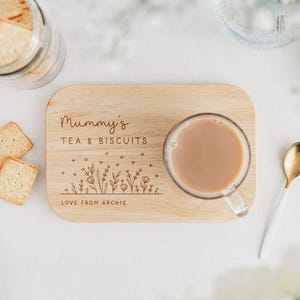May include: A wooden tea and biscuit serving board with the text "Mummy's TEA & BISCUITS" and a floral design. A clear glass mug of tea sits on the board, with biscuits and a gold spoon nearby. The text "LOVE FROM ARCHIE" is also on the board.