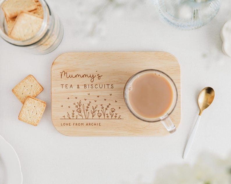 May include: A wooden tea and biscuit serving board with the text "Mummy's TEA & BISCUITS" and a floral design. A clear glass mug of tea sits on the board, with biscuits and a gold spoon nearby. The text "LOVE FROM ARCHIE" is also on the board.