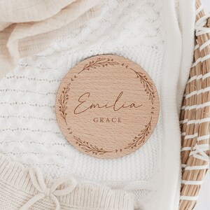 May include: A round wooden plaque with a leafy wreath design and the name "Emilia Grace" engraved in cursive script.