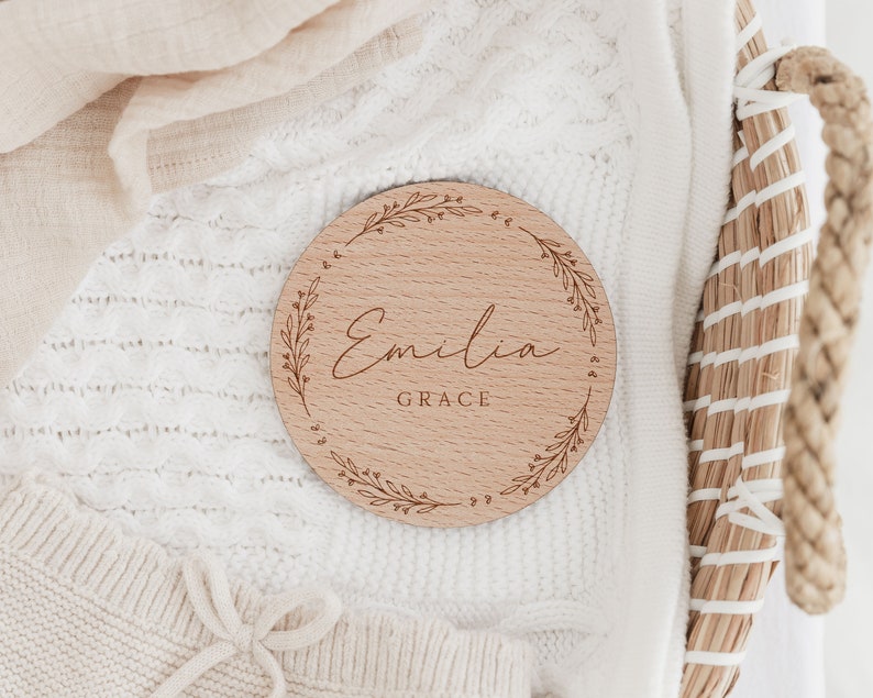 May include: A round wooden plaque with a leafy wreath design and the name "Emilia Grace" engraved in cursive script.