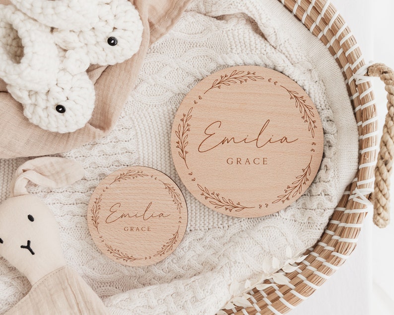 May include: Two wooden plaques with a floral design and the name "Emilia Grace" engraved on them. The plaques are on a white knitted blanket in a wicker basket.